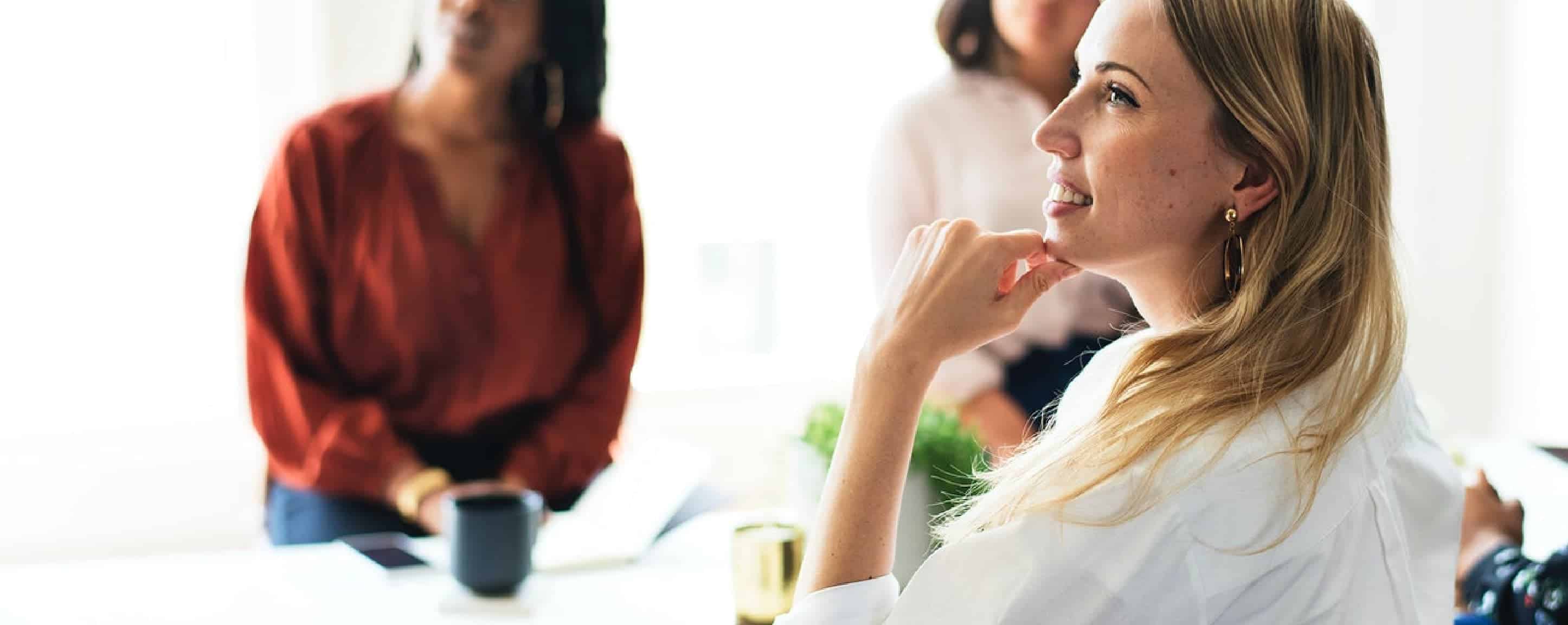 BIZ-LYNX Technology - A woman with blonde hair, dressed in white, sits attentively with her hand on her chin. In the background, two other people, slightly out of focus, sit and listen. Her posture reflects the essence of who we are—dedicated professionals committed to exceptional customer service.