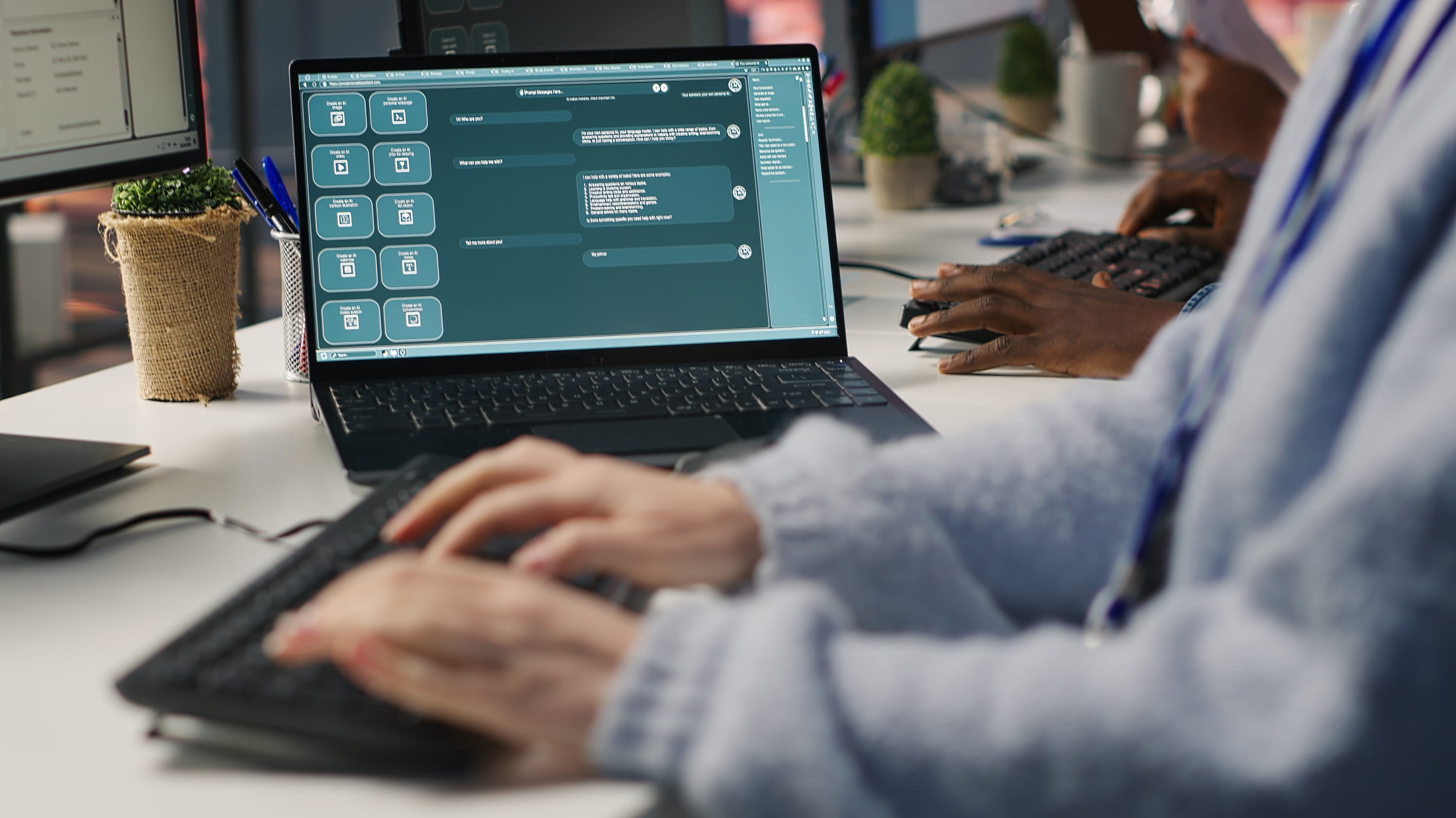 An office worker types on a laptop showing an AI chatbot conversation, with a colleague working at a nearby desk.