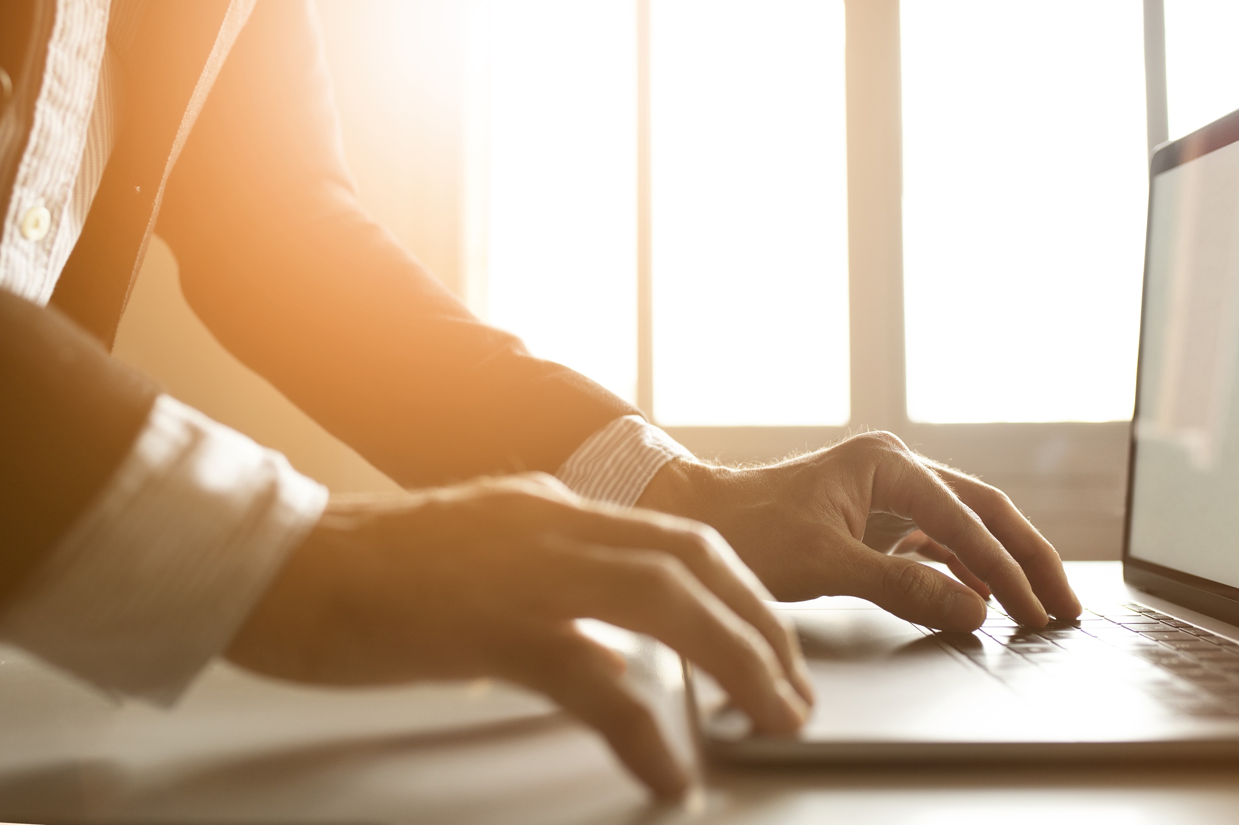 Close-up of a person's hands typing on a laptop keyboard, bathed in warm sunlight through office windows