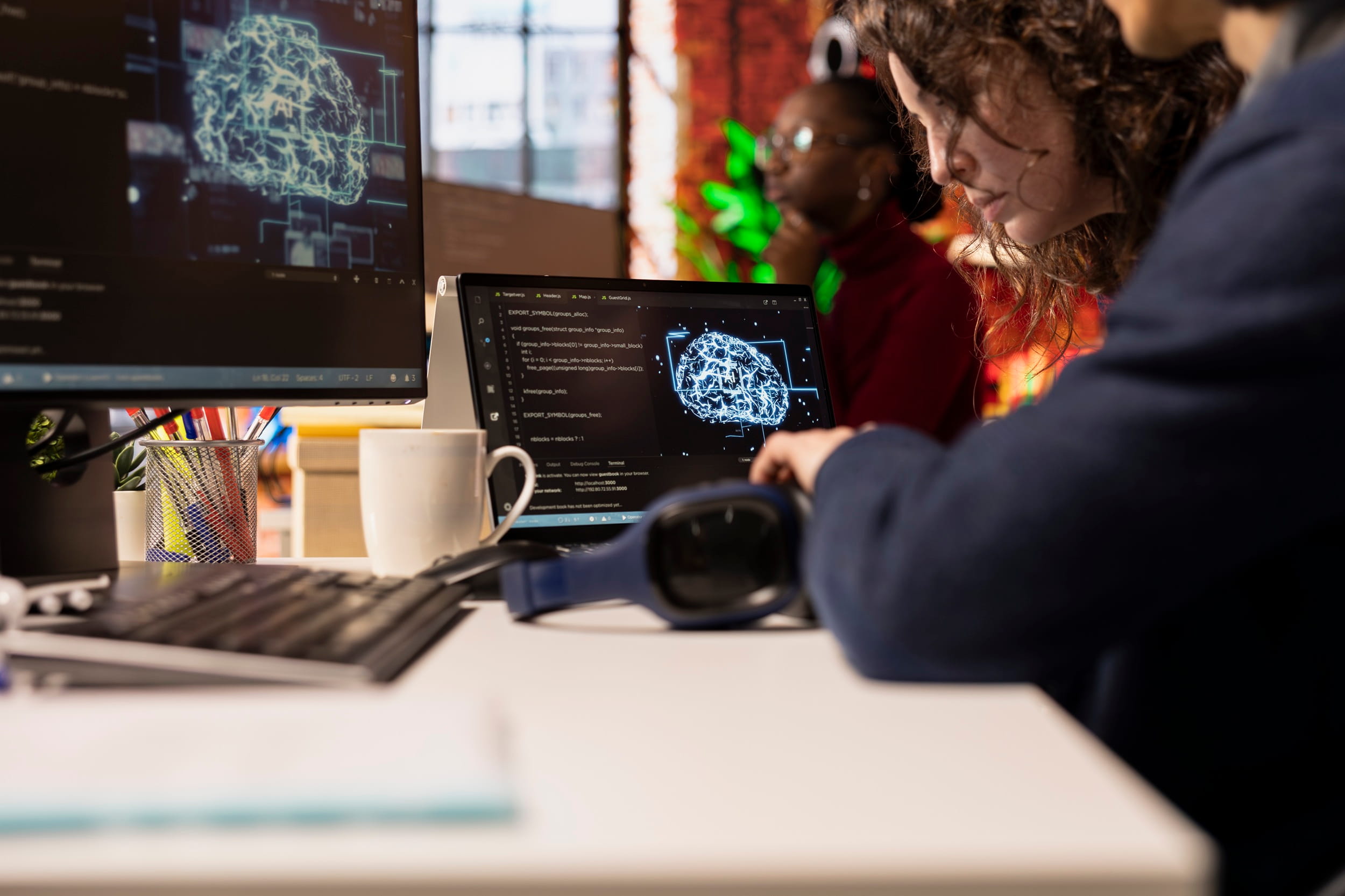 Two IT professionals review a glowing digital brain graphic and code on a monitor and laptop in a casual office.